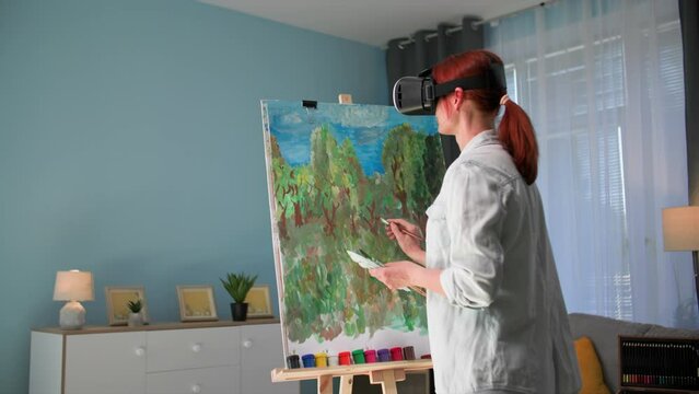 modern art, creative female artist paints a picture on canvas using paints and a brush in viar glasses standing in a cozy room