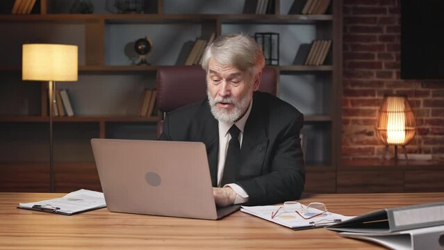 Cheerful senior man in formal clothes looking for correct file while talking with colleague via video call app at work. Smiling general manager telling jokes with attendee during office hours.