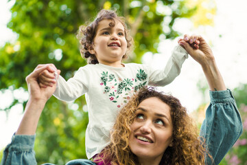 Obraz premium Joyful Moments in Nature: A cheerful young child with curly hair enjoys a playful moment, being lifted up by a loving woman, surrounded by the beauty of lush greenery.