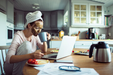 Young woman with a cosmetic mask on her face drinking coffee and doing financials on the laptop in the kitchen at home