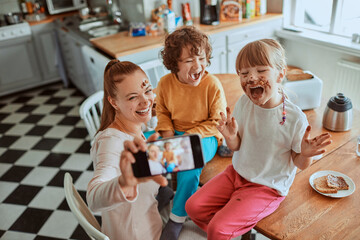 Young mother taking a selfie with her children on a smartphone while eating breakfast and being messy in the kitchen - Powered by Adobe
