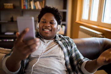 Young man listening to music on the headphones while using a smartphone on the couch at home