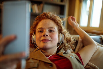 Young redhead woman listening to music on the headphones while using a smartphone on the couch at home