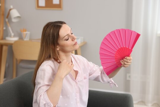 Woman waving pink hand fan to cool herself on sofa at home
