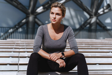 Woman athlete wearing female sportswear sitting on bench of bleachers in outdoor stadium