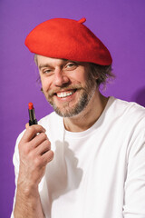 Funny middle aged man wearing red beret is holding lipstick in his hand against purple background