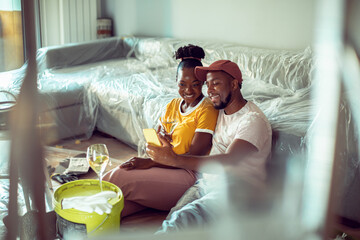 Young African American couple taking a selfie on smartphone while drinking wine on a break from painting their living room at home