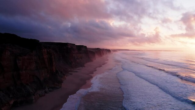 Inspiring and cinematic pan out drone shot of incredible cliffs going into ocean. Portuguese beach during epic pink sunset. Romantic and dreamy travel wanderlust mood