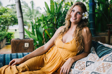 Joyful adult woman with flower in hair dreaming while resting on sofa in tropical garden during sunny day