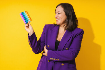 Cheerful middle aged woman wearing purple blazer playing pop-it fidget toy against yellow background