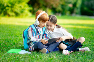 Naklejka na ściany i meble a boy and a girl with backpacks are sitting on the lawn and reading a book and a tablet with headphones, the concept is back to school, brother and sister are doing homework on the street