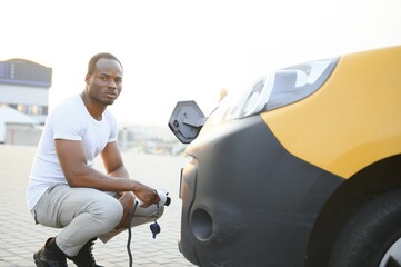 african man holding charge cable in on hand standing near electric car.