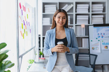 Successful Asian businesswoman smiling using laptop computer and holding coffee cup at office. Confident Asia businesswoman sitting happily in the office.