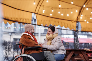 Obraz na płótnie Canvas Side view portrait of smiling young woman holding hands with man in wheelchair enjoying date outdoors in winter