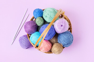 Lots of colorful knitting balls in a basket with knitting needles on a pastel purple background....