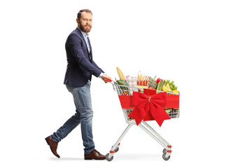 Bearded young man walking and pushing a shopping cart with food and tied with a red ribbon