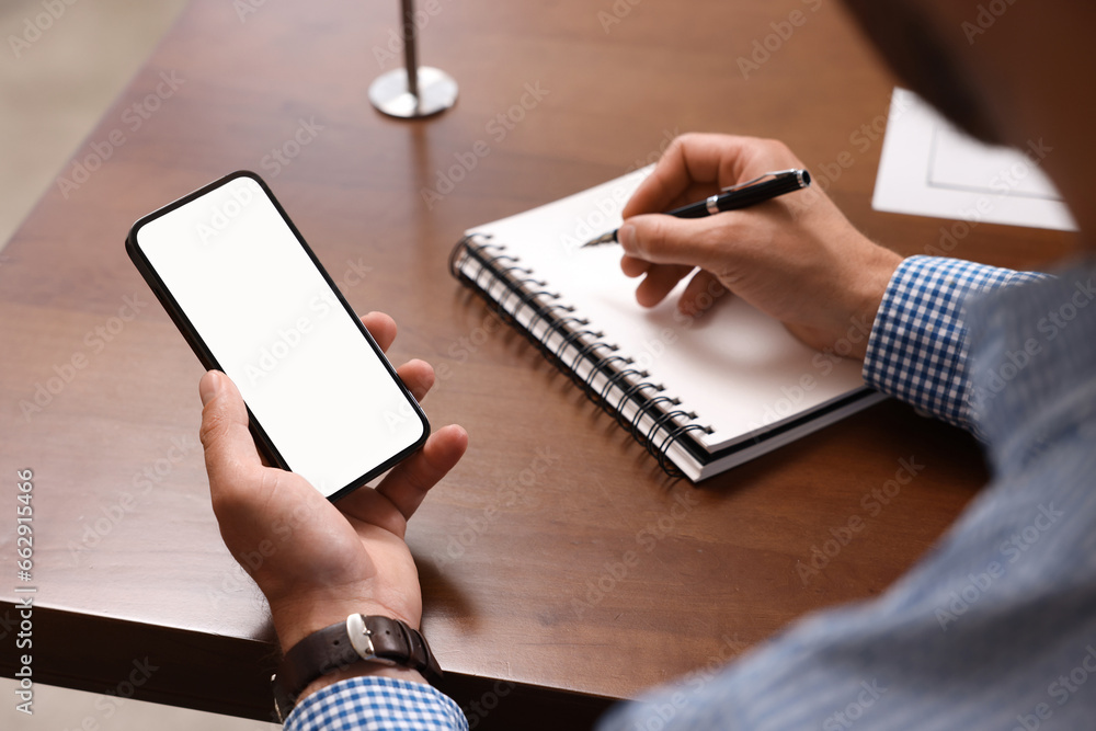 Poster man with smartphone writing something in notebook at table, closeup