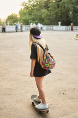 Photo from the back of a young blonde girl in black clothes and a cap with a backpack on her shoulders, riding a skateboard.