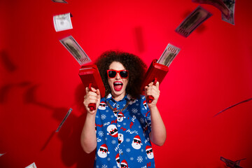 Photo of shocked cheerful woman wear ugly santa claus print x-mas shirt shooting cash flying gun isolated red color background