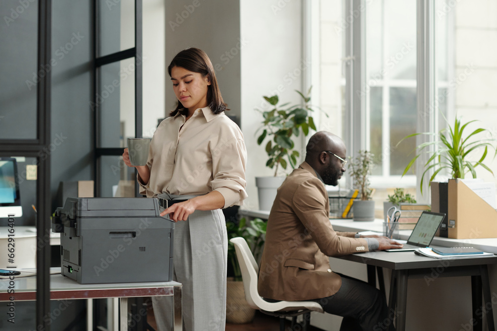 Aufkleber young businesswoman with cup of coffee pressing button on copier panel while standing by xerox machi