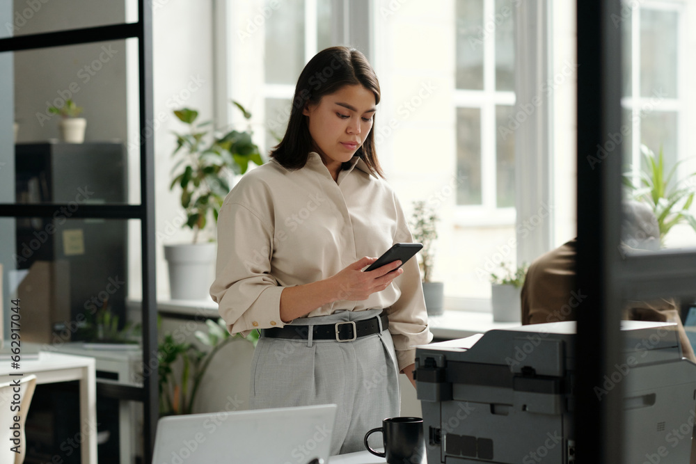 Poster young pretty female office manager with mobile phone texting by workplace while standing in front of