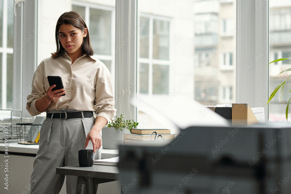 Poster young secretary with dark hair looking at documents in printer and using smartphone while standing a
