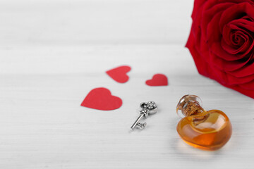 Heart shaped bottle of love potion with small key, paper hearts and red rose on white wooden table,...