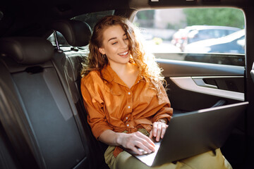 A young curly woman with a laptop sitting in the back seat of a car. Beautiful woman uses laptop in car. Remote work, freelance.
