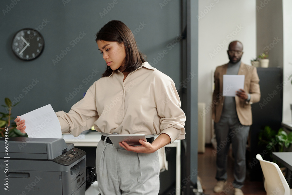 Poster young serious businesswoman with tablet taking printed paper document from xerox machine while stand