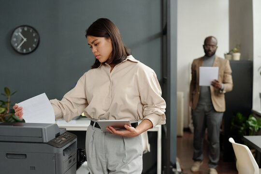 Young serious businesswoman with tablet taking printed paper document from xerox machine while standing against coworker and using tablet