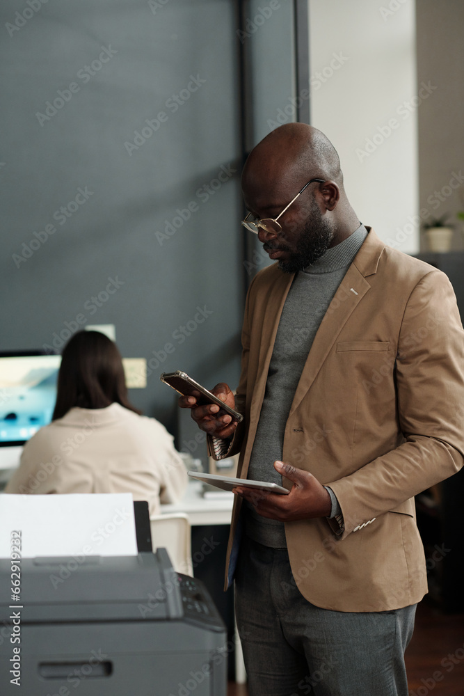 Poster young african american businessman using smartphone and tablet while standing in front of xerox mach