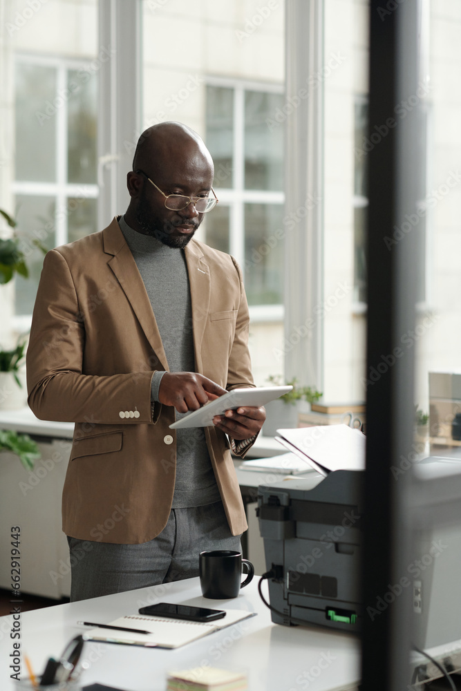 Poster Young confident businessman in formalwear pointing at tablet screen while networking in front of xerox machine with copies of paper documents