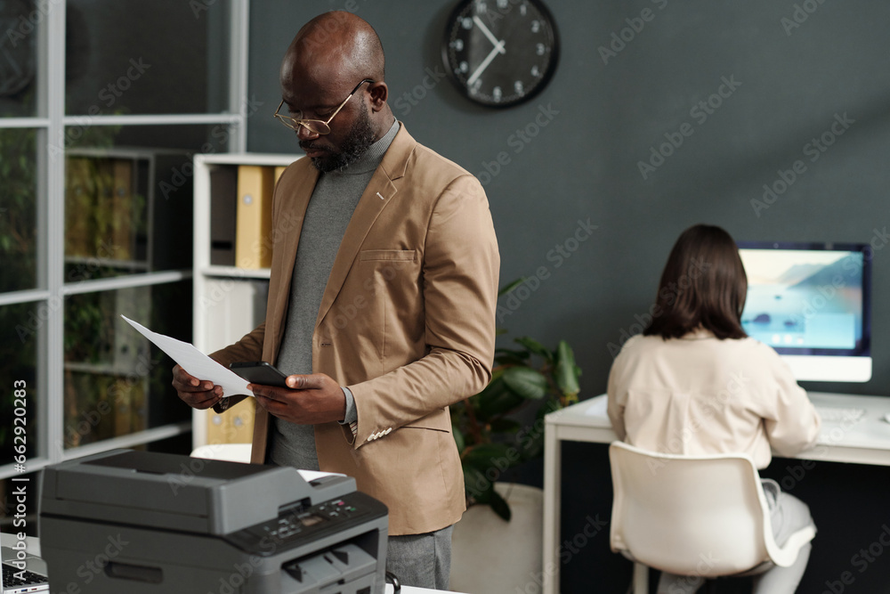 Poster young serious african american ceo comparing data in smartphone with that on paper document while st