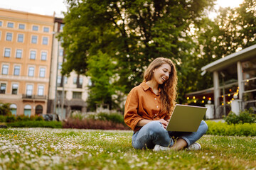 Joyful woman with laptop outdoors. A young woman sits on a green lawn and works with a laptop. Concept of blogging, freelancing, relax.