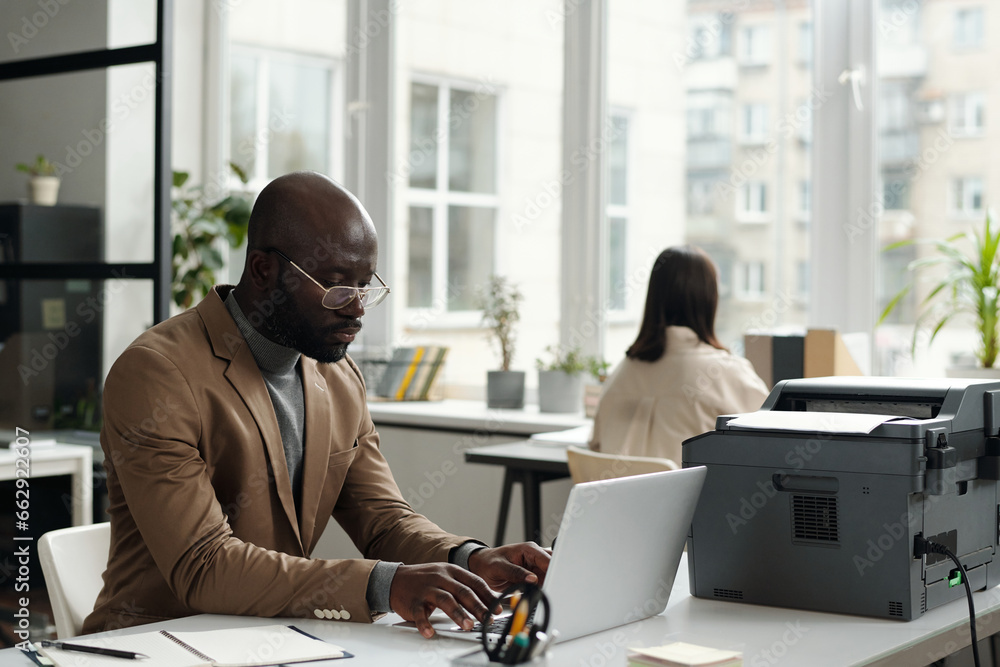 Aufkleber young serious white collar worker typing on laptop keyboard while sitting by workplace with xerox ma