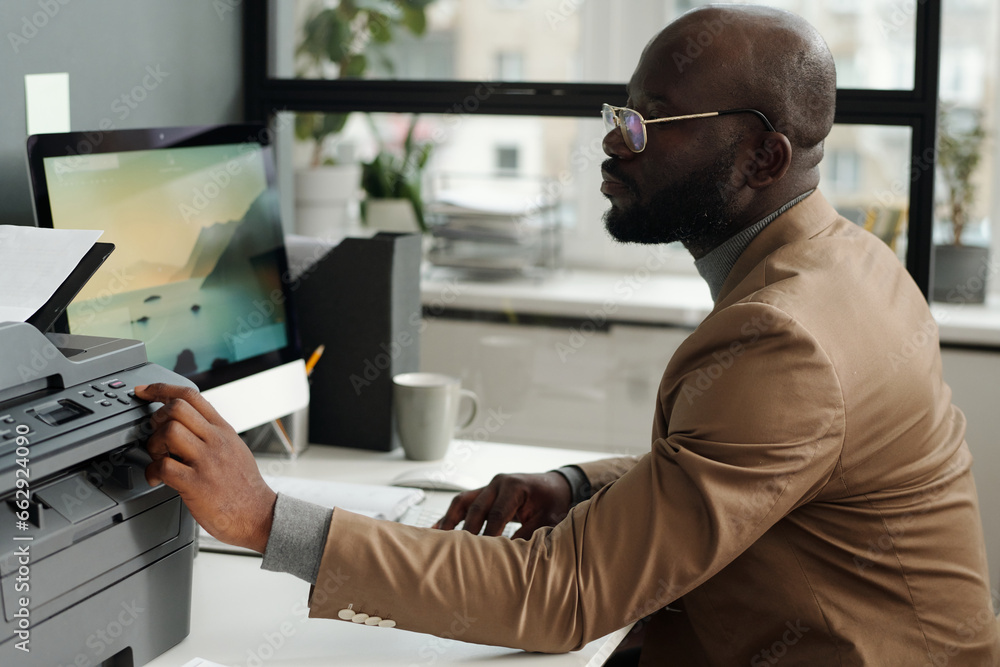 Fototapete Young businessman in eyeglasses and formalwear pressing start button on panel of printer while sitting by workplace in front of computer screen