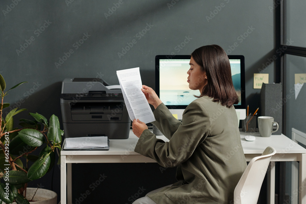 Poster Side view of young secretary or accountant looking through financial documents while sitting by her workplace in front of computer