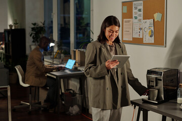 Young successful businesswoman looking at tablet screen and making cup of coffee while watching online video or scrolling through news