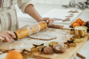 Woman making christmas gingerbread cookies in modern white kitchen close up. Hands kneading gingerbread dough with rolling pin, golden metal cutters, cooking spices, festive decorations