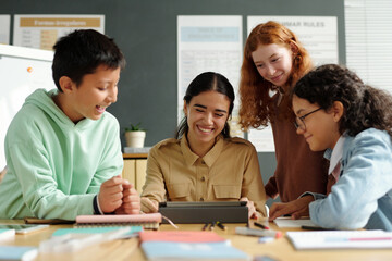 Young cheerful teacher and group of pupils looking at tablet screen and discussing new rules of English grammar at lesson or language courses