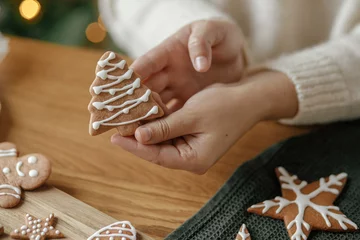 Foto op Plexiglas Hands holding gingerbread cookie tree with icing on rustic wooden table. Atmospheric Christmas holiday traditions, family time. Woman decorating cookies with sugar frosting © sonyachny