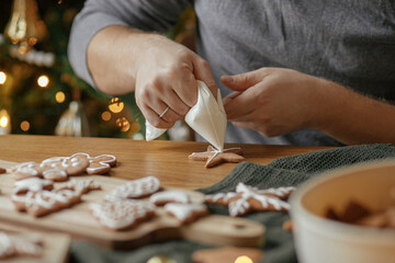 Decorating gingerbread cookies with icing on rustic wooden table at christmas tree golden lights. Atmospheric Christmas holiday traditions. Man decorating cookies with sugar frosting
