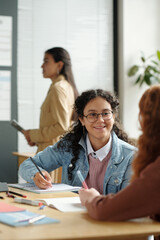 Obraz premium Cute smiling schoolgirl with pen and copybook looking at classmate at lesson of English language during discussion of new grammar rules