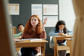 Clever schoolgirl raising hand while sitting by desk and looking at whiteboard during discussion of new rules of English grammar