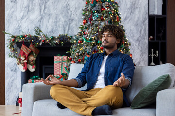 Christmas young man meditating in lotus pose sitting on sofa in living room, hispanic man doing breathing exercises calm down and relaxing new year celebration. - obrazy, fototapety, plakaty