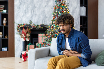 New Year's celebration and Christmas, man has severe stomach pain, hispanic sitting on sofa in living room, massaging stomach with hand. - obrazy, fototapety, plakaty
