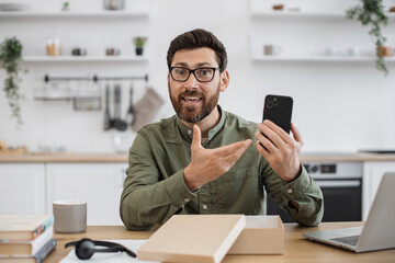 Mobile web camera view of technology blogger making review of modern smartphone during video filming at cozy home. Caucasian bearded man satisfied online purchase during holiday discount.