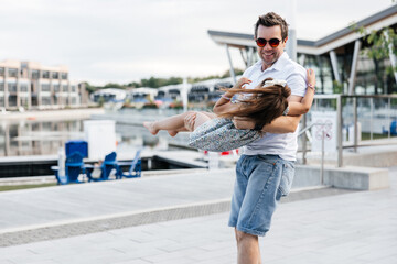 Fototapeta premium A happy father holds his daughter in his arms and spins her around. Dad and daughter are having fun walking outside. Happy fatherhood and childhood. Dad and his little daughter having fun