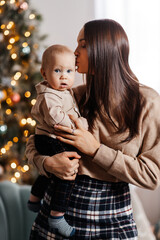 Mom holds her little son in her arms against the backdrop of a decorated Christmas tree. A young mother and her little son are celebrating Christmas. Vertical portrait of a mother and her son