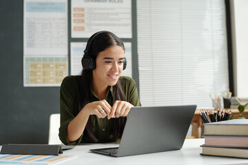 Young smiling teacher or student of English language course in headphones sitting by desk in front...